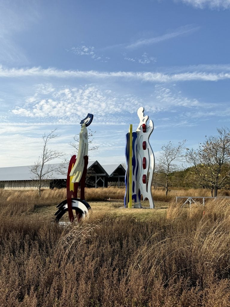 In a field, a pair of brightly colored sculptures in the form of brush strokes by Roy Lichtenstein