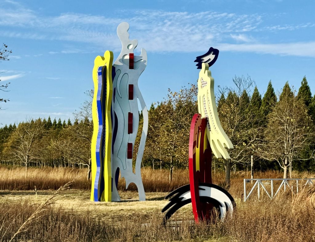 In a field, a pair of brightly colored sculptures in the form of brush strokes by Roy Lichtenstein