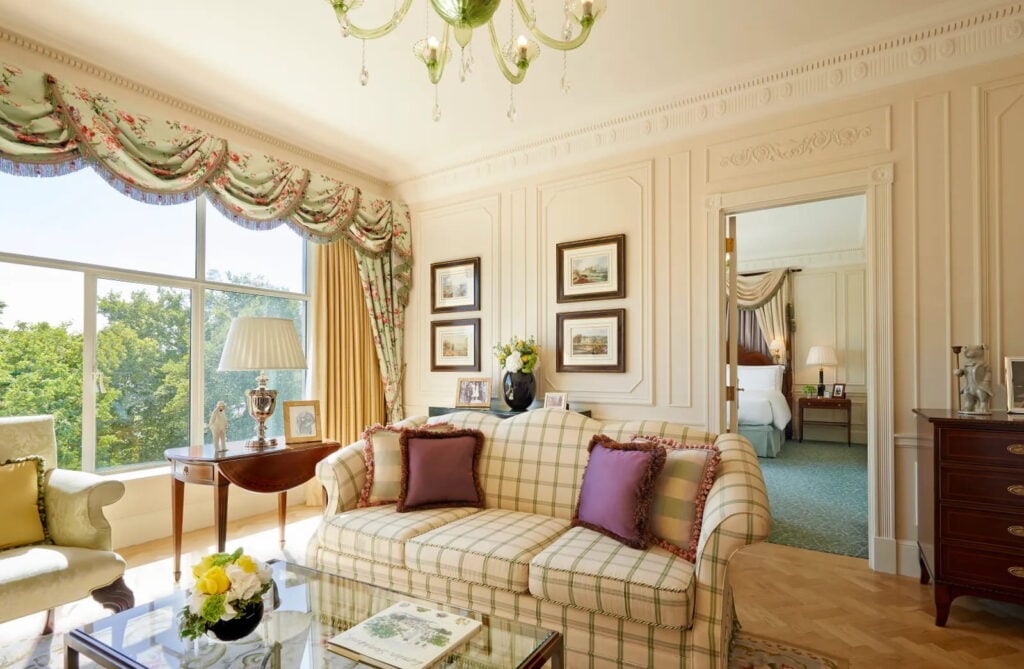 Photograph of a brightly lit hotel suite with a large comfy couch with purple pillows surrounded by tables, chairs, and pictures on the walls underneath a little chandelier next to a large window