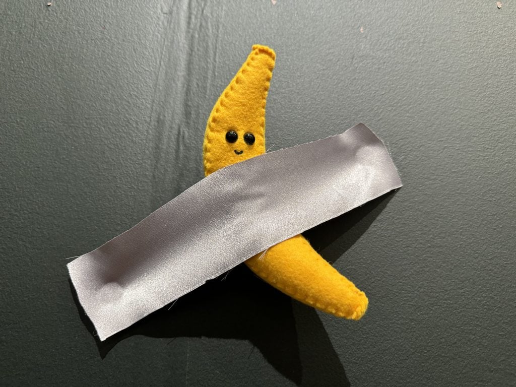 Lucy Sparrow's $250 limited edition Maurizio Cattelan-inspired banana at the "Blessed Be the Fruit" installation at the SCOPE Art Show in Miami Beach. It is a felt stuffed banana with black plastic eyes and a tiny grin affixed to a black wall with gray fabric that looks like duct tape. 