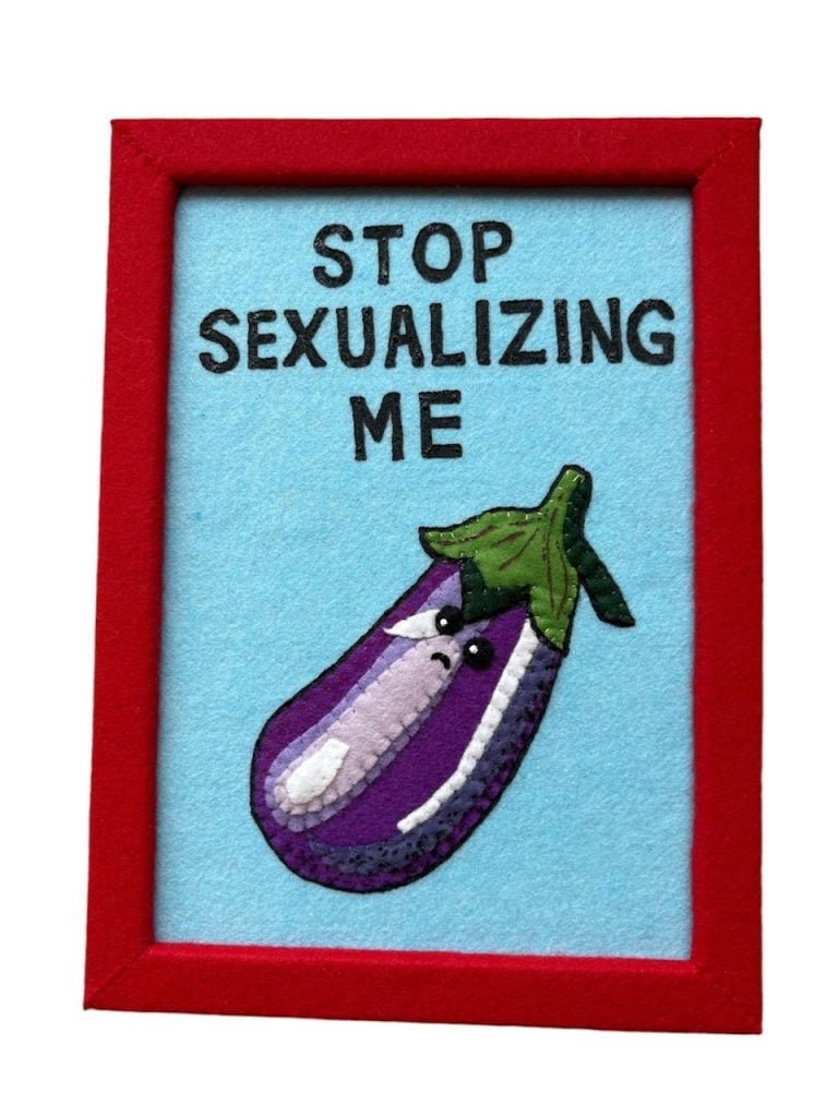 Lucy Sparrow's eggplant from her "Family Portrait" series. It is a flat, framed felt artwork of a sad eggplant that says "stop sexualizing me."