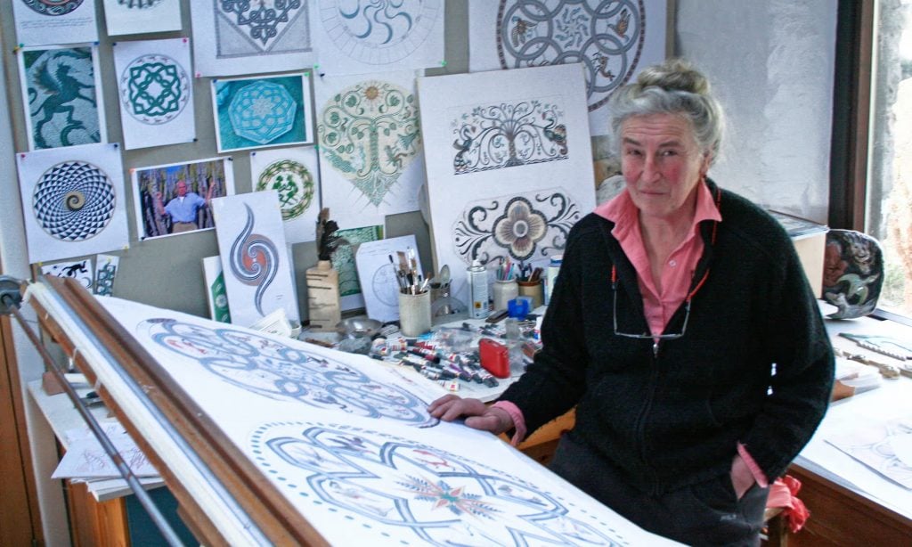 Maggy Howarth in her studio. She is an elderly woman with long gray hair in a bun on the top of her head, sitting at a drafting table surrounded by preparatory drawings for her pebble mosaics. 