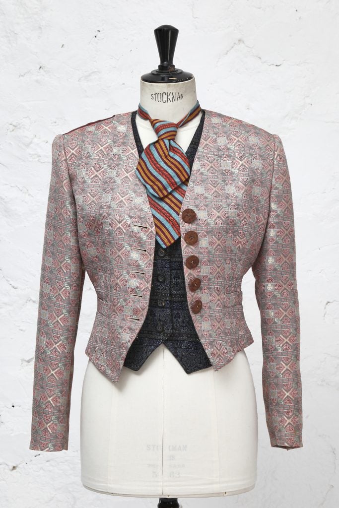 A photograph of a patterned jacket atop a dark waistcoat with a striped neck tie on a white mannequin depicted before a white background.