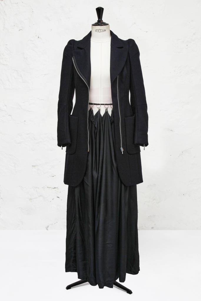 A photograph of a mannquin draped with a long black jacket and a black skirt in the style of hanging from a waistband on eyelets like a curtain, all depicted against an all-white background.