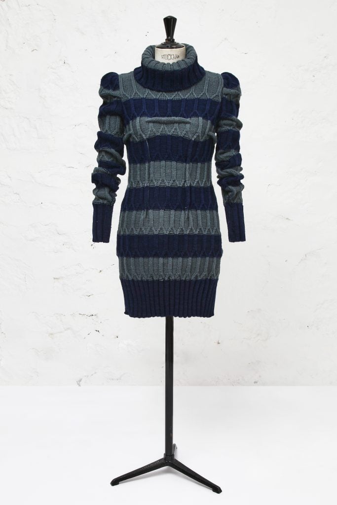 A photograph depicting a blue and gray striped sweater dress with long ruched sleeves on a stand before an entirely white background.