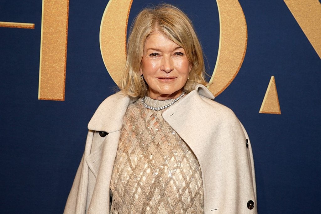 A photograph of Martha Stewart wearing a beige jacket over a gold, patterned shirt, before a blue wall with gold lettering on it.