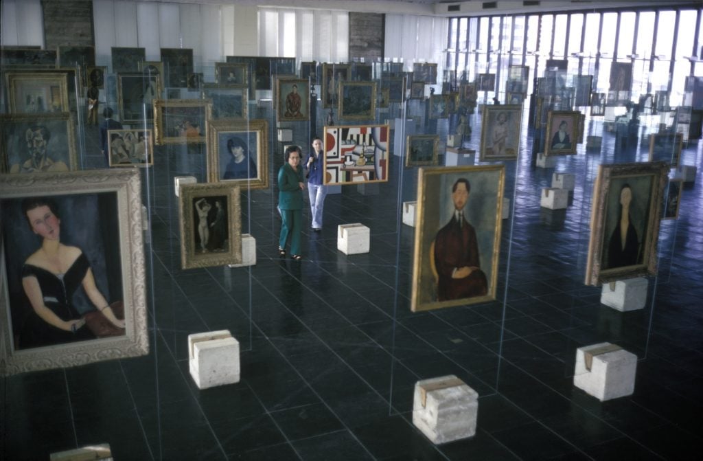 Art gallery featuring numerous framed paintings displayed on transparent glass easels, creating a floating effect. The room has a grid-like floor and large windows, with two individuals viewing the artwork in the foreground