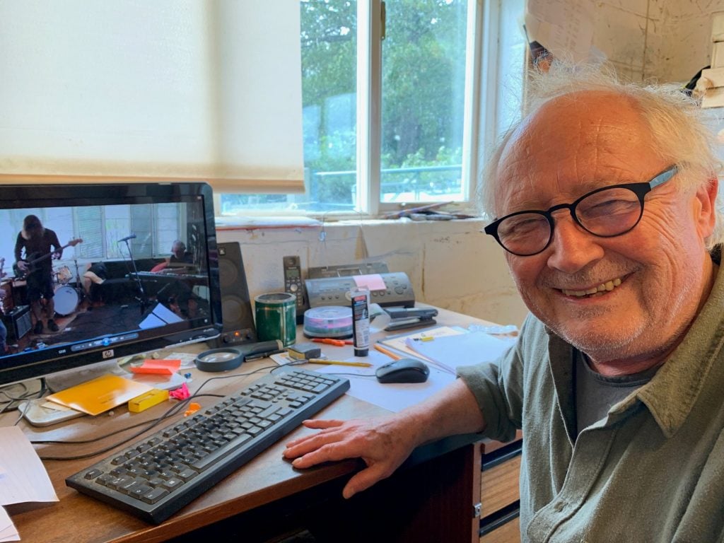 Mel Ramsden in the studio (2019). He is an elderly bald white man in black rimmed glasses, smiling widely and sitting at his desk in front of a keyboard and computer, in front of a window. 