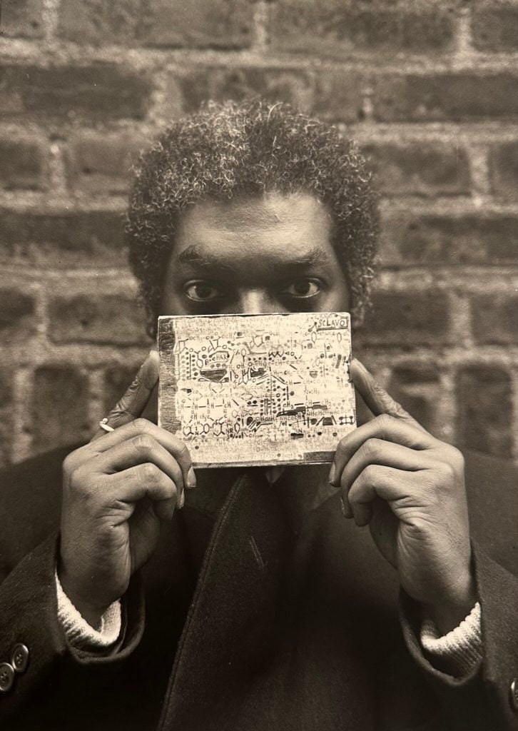 A sepia toned photo of Melvin Way, a young Black man with graying hair, holding one of his drawings in front of his nose and mouth, standing against a brick wall, in 1994. 