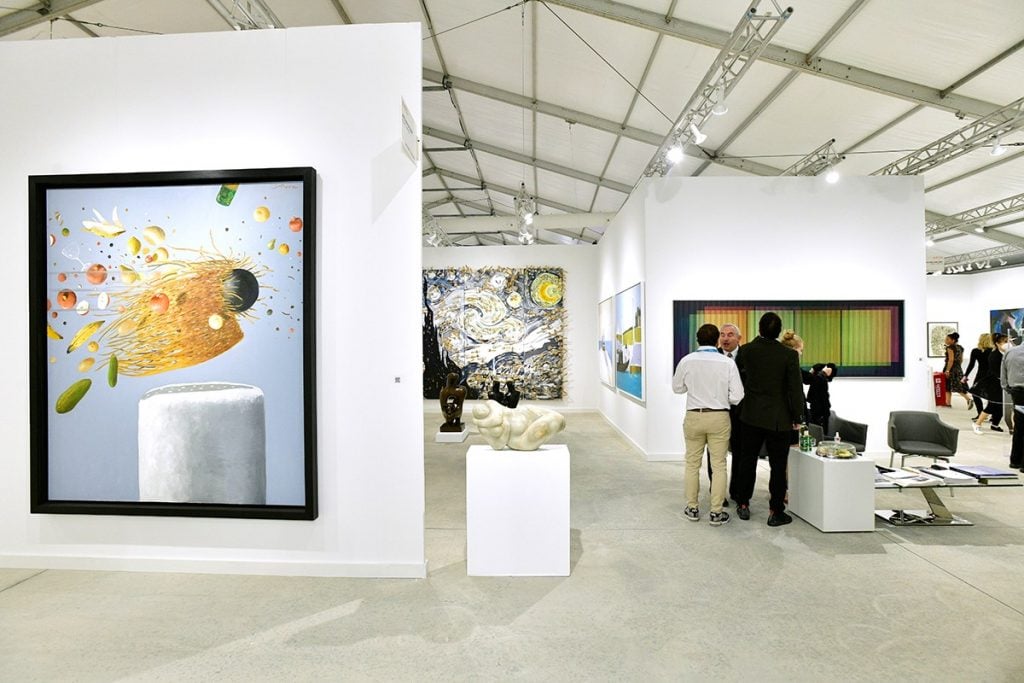 "Interior view of an art gallery showcasing diverse artworks, including vibrant paintings, sculptures, and attendees engaging in conversation under a bright, modern setting.