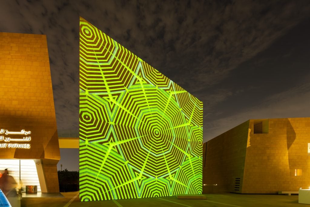 Exterior of Diriyah Art Futures showing a projection mapping illuminates a modern building's facade at night with vibrant green geometric patterns, creating a dynamic and visually striking architectural display.