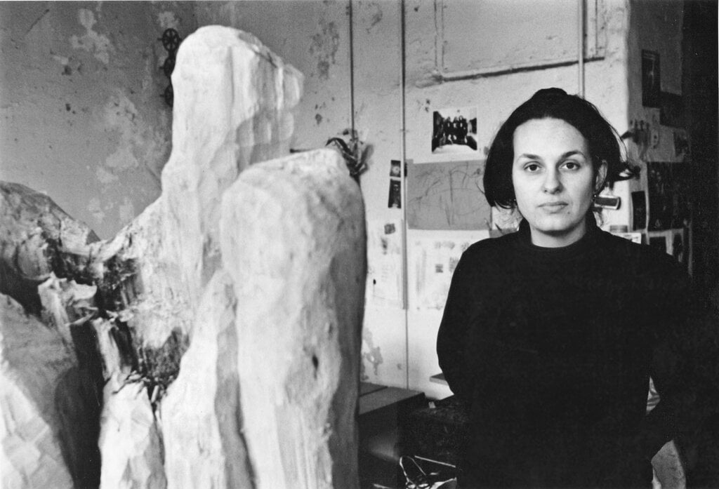 A black and white photo of young white artist Nancy Azara in her studio in the 1970s, seen from the waist up next to a monumental, roughly carved wooden sculpture. . 