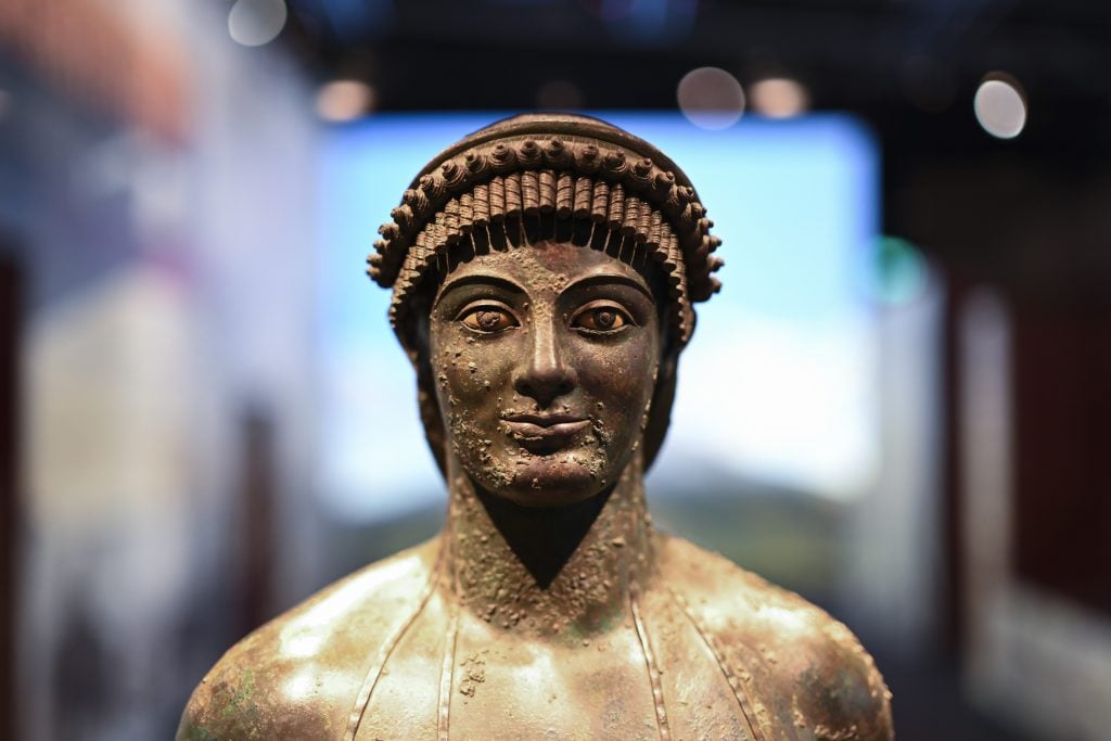 A close-up of an ancient bronze statue’s face, with intricate headgear details.