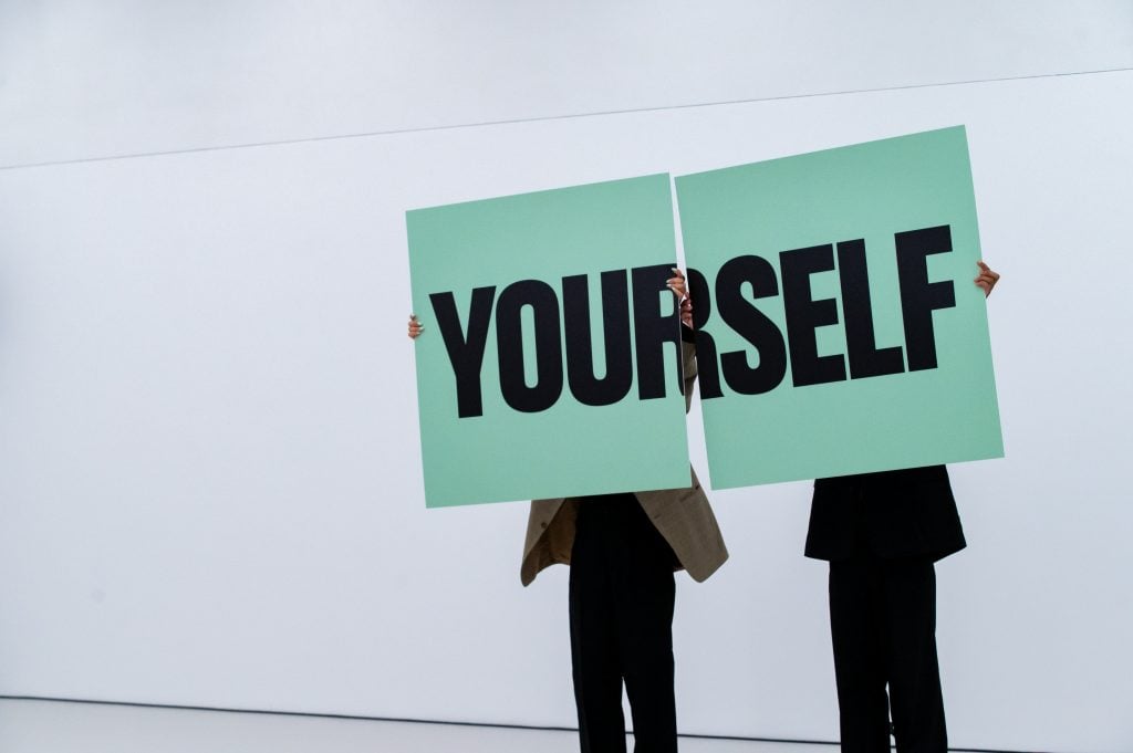 A man and woman holding up giant green placards that join together to read "YOURSELF"