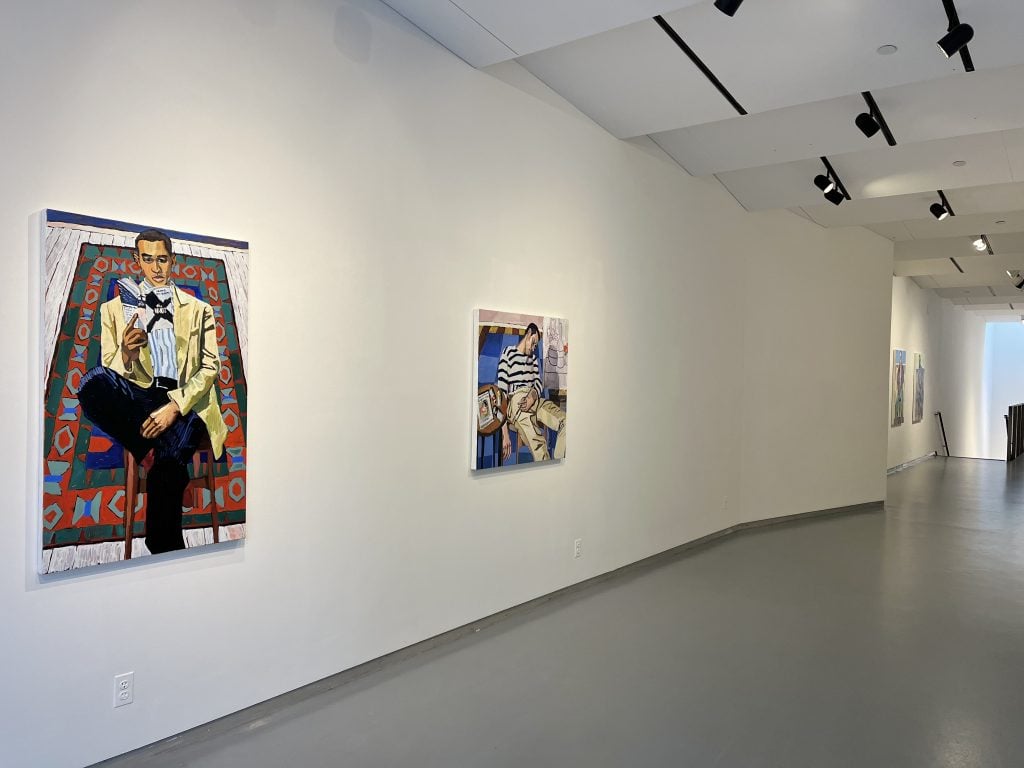 A photograph of two figurative paintings by Nicolas Coleman hanging on a white wall at Thierry Goldberg above a gray floor benearth track lighting, in a passageway heading towards a set of descending stairs.