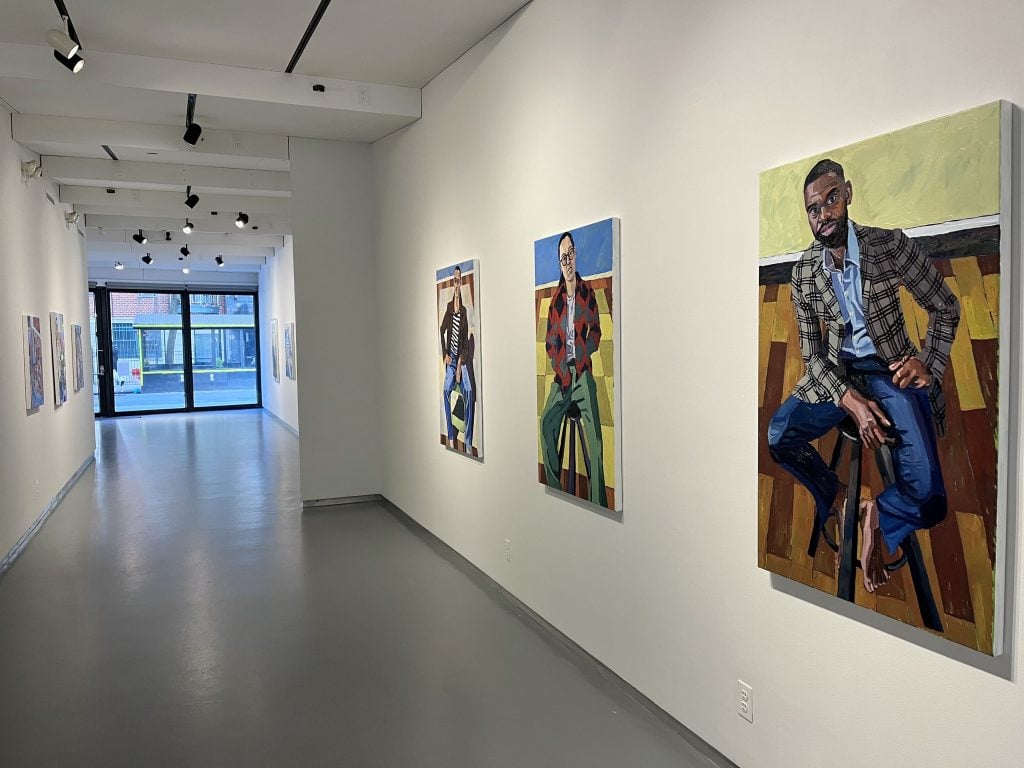 A photograph of two figurative paintings by Nicolas Coleman hanging on a white wall at Thierry Goldberg above a gray floor benearth track lighting, in a passageway heading towards glass doors leading outside.