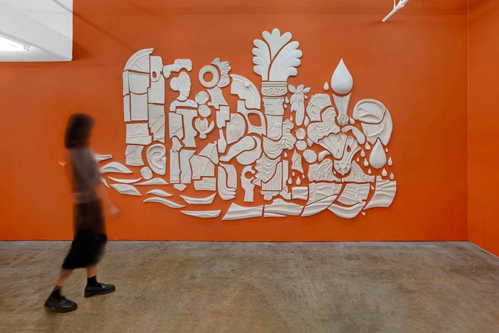 Nina Surel, Allegory of Florida. A slightly blurry woman is photographed in motion in front of a large white ceramic wall relief with 100 separate pieces, hung on a bright orange wall. 