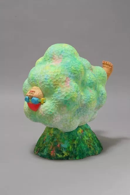 Playful sculpture of a human-like figure resembling a fluffy green cloud, wearing colorful sunglasses, with a foot sticking out, set on a textured base.