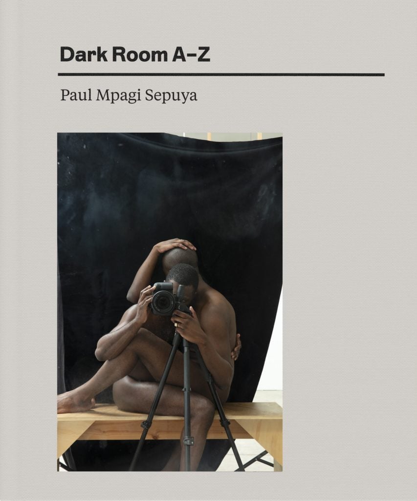 The⁢ cover for Paul Mpagi Sepuya's Dark ​Room A-Z is picture with a photograph ‍of a nude Black man on a ⁤table in ⁤a photo studio