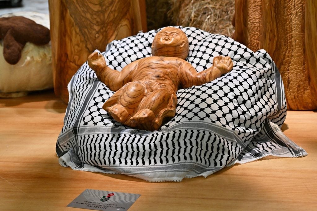 a wooden figure of a baby is resting on a mound that is draped in a Keffiyeh, a cloth with a simple pattern in black and white