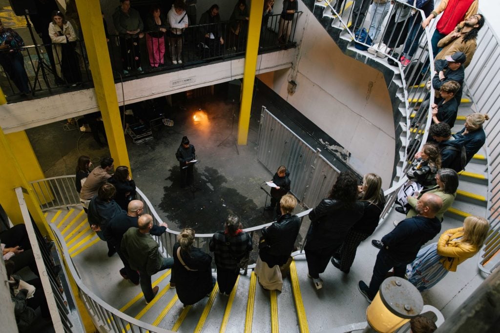 A crowd surrounds a circular staircase as a performer delivers a reading in an industrial space.