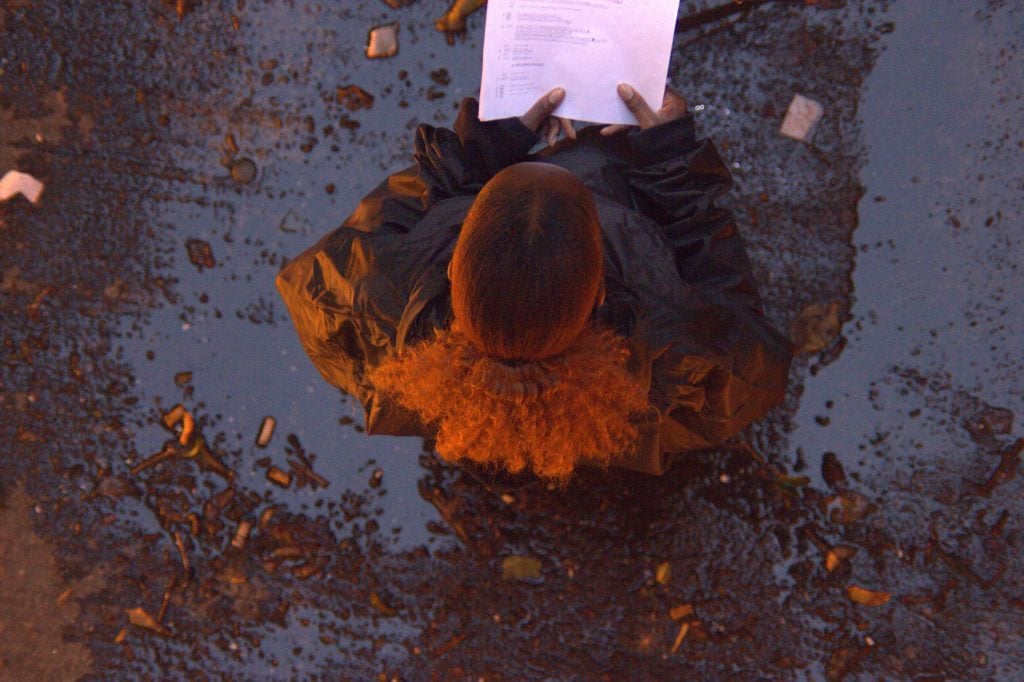 Overhead view of a performer with fiery orange hair reading from a paper, reflections on wet ground.