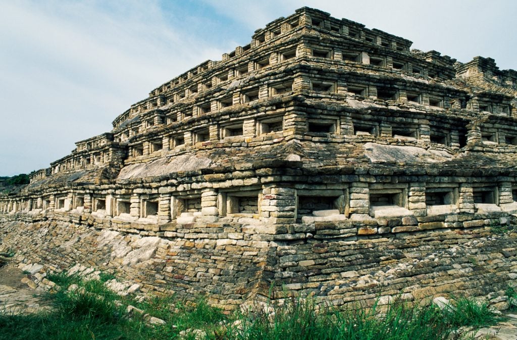 a photograph of a large latin american pyramid made from old scarred grey stone with niches and notches carved into the exterior steps in symmetrical pattern