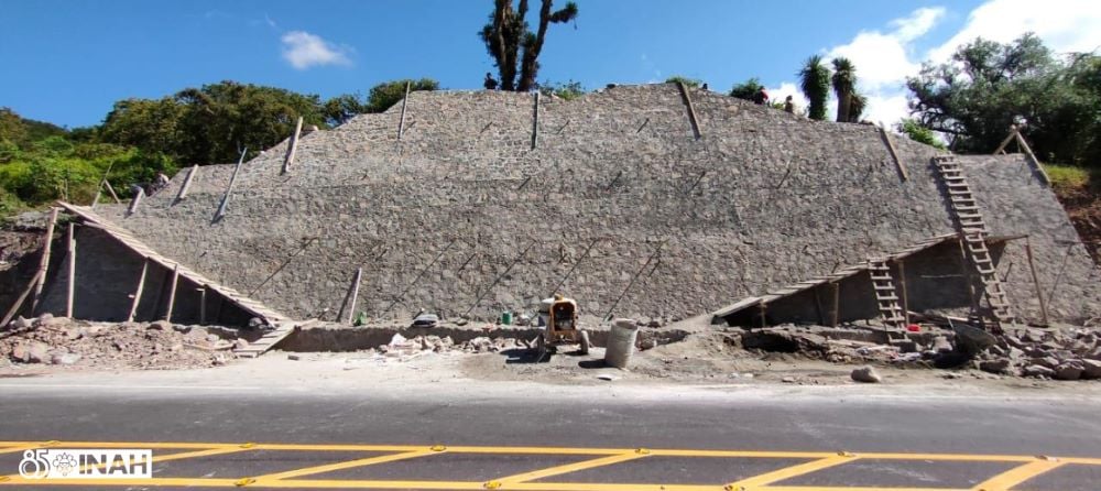 photograph of a large partially excavated pyramid with small staircases on the side in front of a road under construction with lush trees and a blue sky in the background