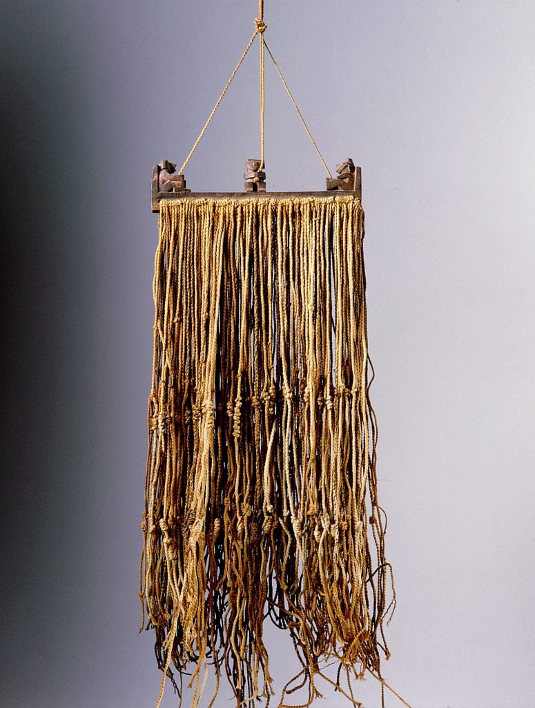 photograph of a collection of weathered brown yellowish strings tied to each other and suspended by a tiny little string from the ceiling against a gray background that goes from dark to light