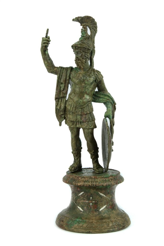 An ancient statuette of Mars in full armor, his right index finger pointing upward