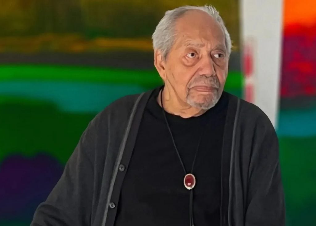 Richard Mayhew, a elderly light-skinned Black man in a black cardigan with a round pendant necklace stands in front of two abstract paintings of hazy bands of color. 