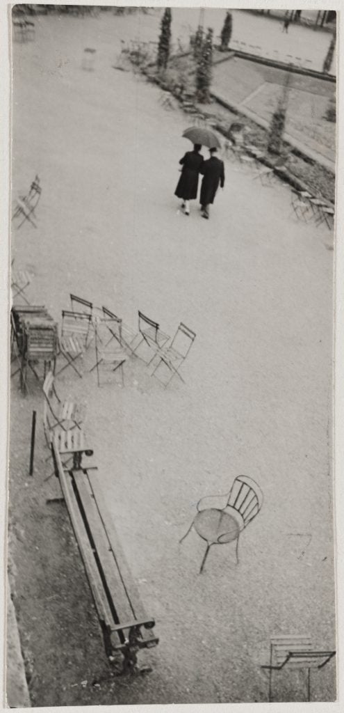 A black and white photograph depicting an empty, snowy park speckled with chairs and one lone couple, as seen from above.