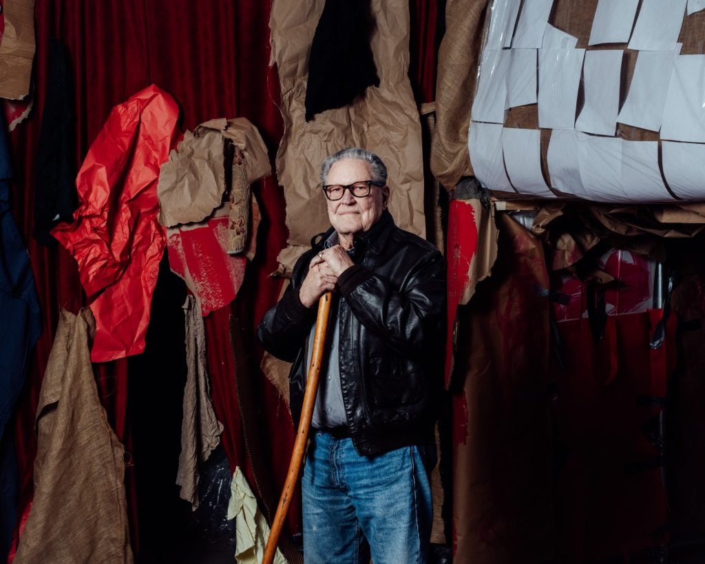 Portrait of Robert Whitman, an elderly white man with gray hair, thick rimmed, glasses, and a black leather jacket and blue jeans, standing with a wooden walking stick in front of crumbled paper. 