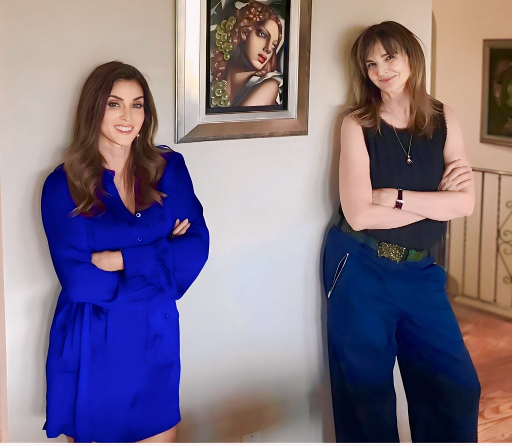 A photograph of two sharply dressed women standing with their arms crossed against a white wall with a small painting hanging between them.