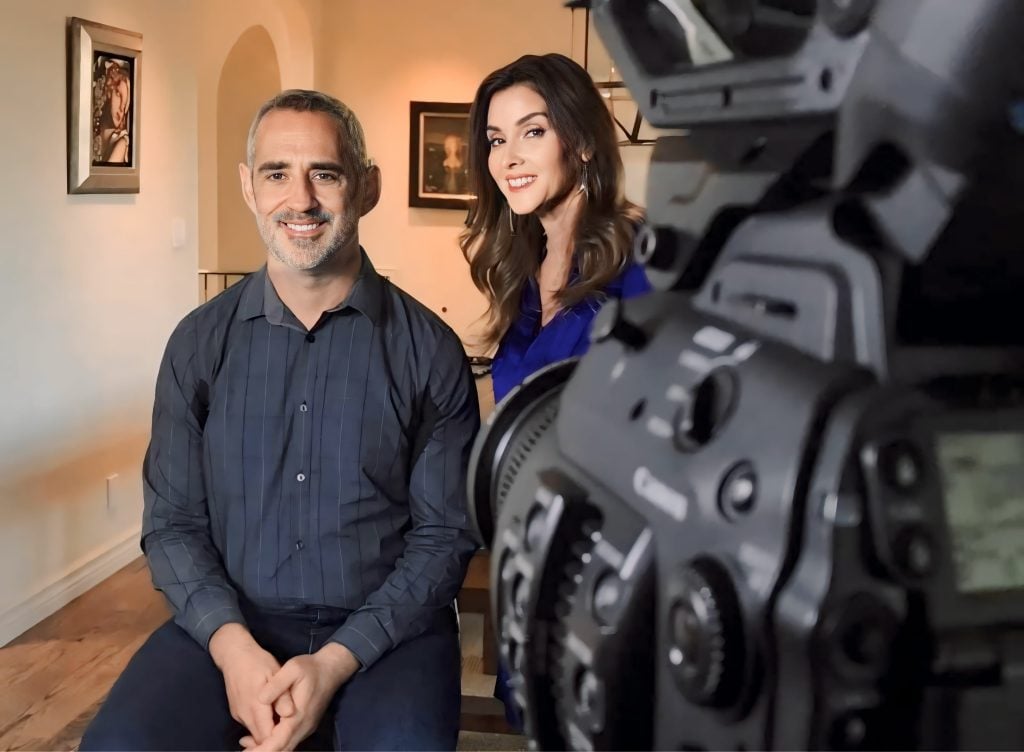 A photograph of a man and a woman smiling side by side on the other end of a camera's lens, inside a nice, warm-lit house.