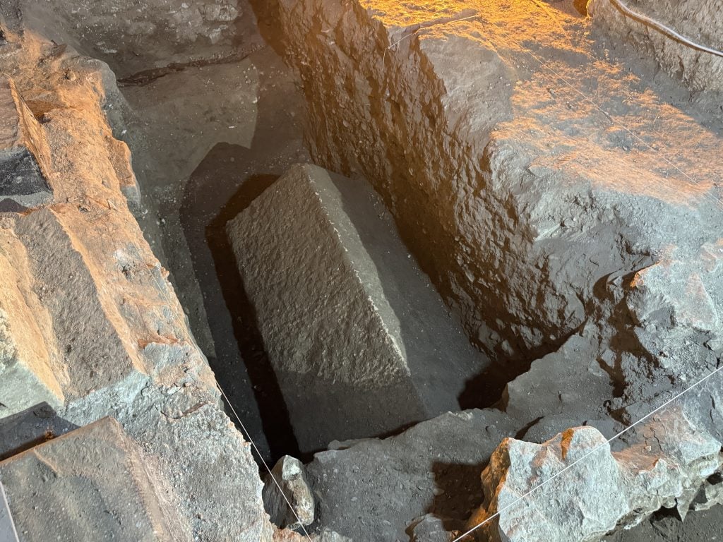 An archaeological excavation revealing an ancient stone sarcophagus partially buried in the ground.