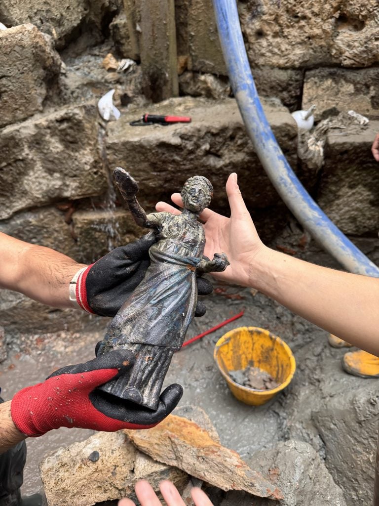 Two pairs of hands holding up a muddied statuette of a goddess