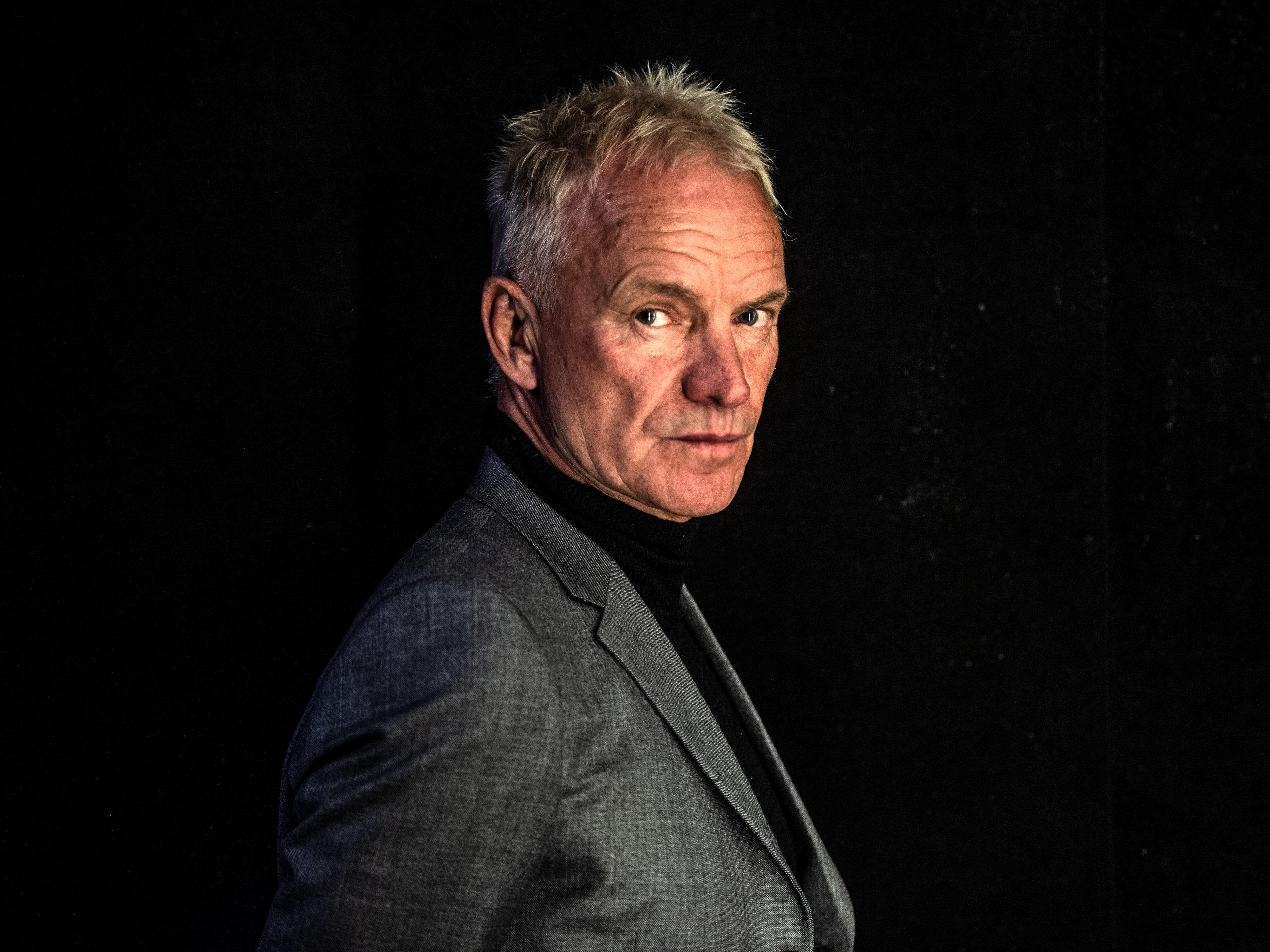 Musician Sting with short greying hair posing in a black top and grey blazer against a black background