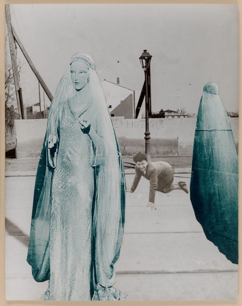 An image of a surrealist collage artwork featuring two blue statues surrounding a black and white picture of a crawling woman, all taking place outside.