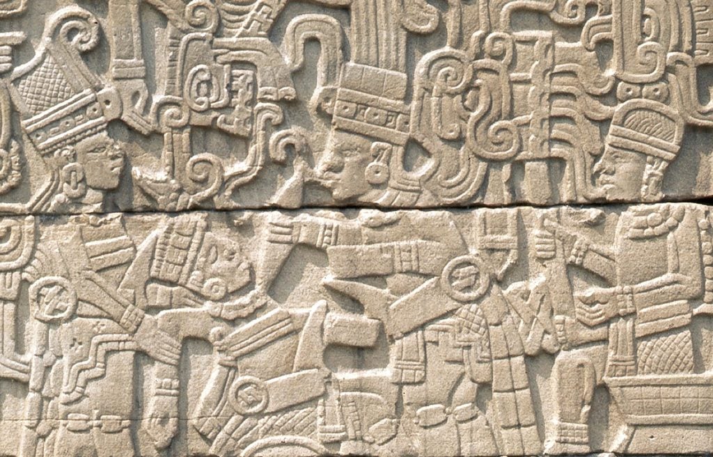 photograph of an engraved relief on a light brown stone wall featuring people in unique headdresses and clothing interacting with each other