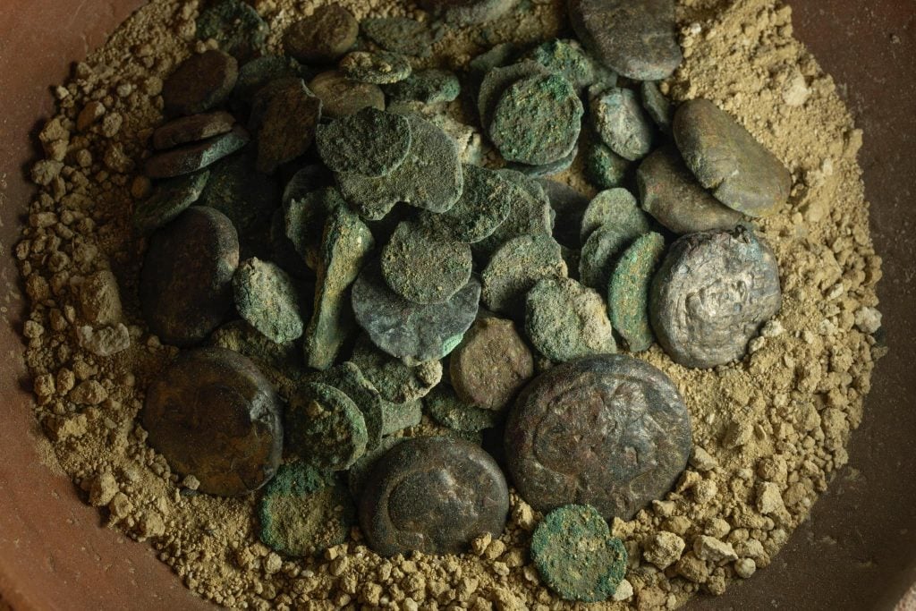 A photograph depicting a hoard of ancient, green-crusted metal coins featuring the undetailed silhouette of a woman in an apparently ceramic bowl.