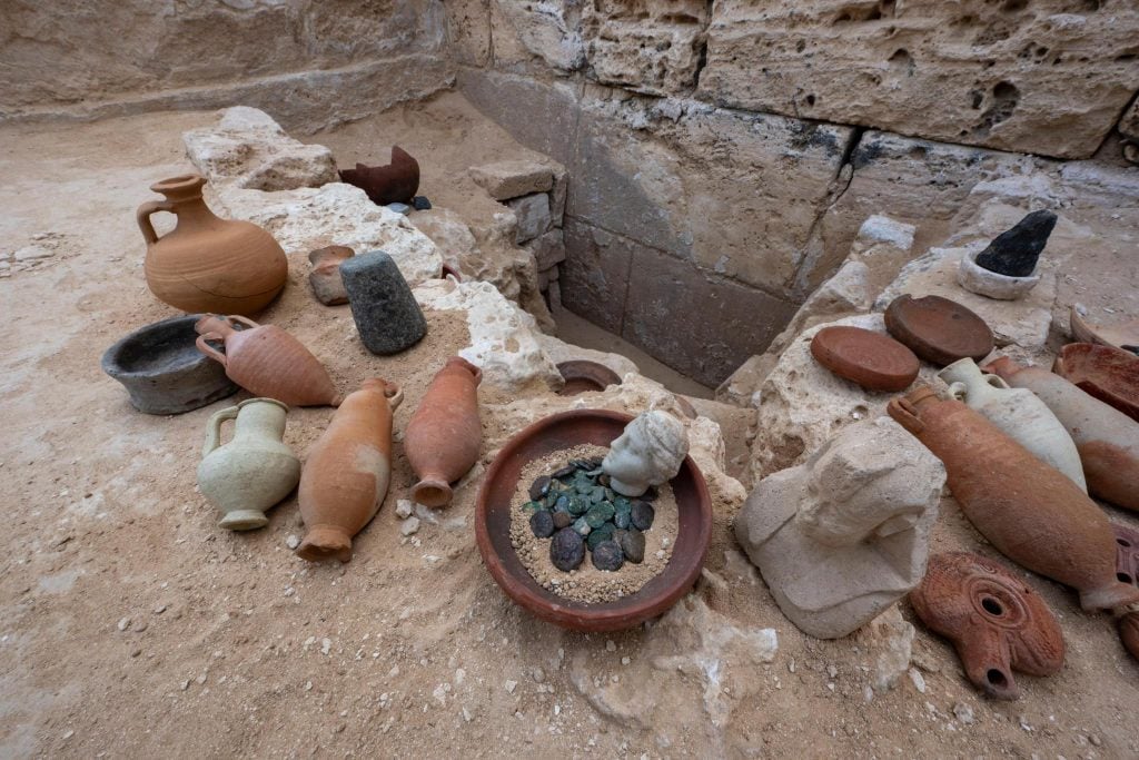 A photograph depicting an array of archaeological artifacts arranged on light brown dirt ground before the hole they were dug up from.