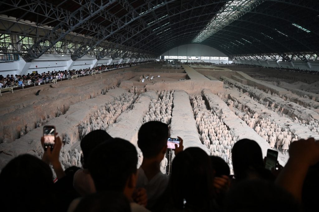 Photograph of a group of people bathed in shadow raising their phones to catch a glimpse of a giant formation of stone warriors inside a large domed room