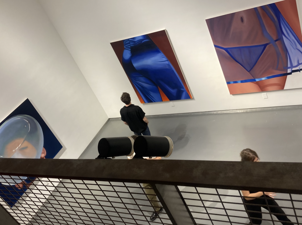 A photograph of several realistic paintings of women's bodies clad in view hanging in the lower level of a white-walled gallery, with a metal railing visible in the foreground and a smattering of viewers below.