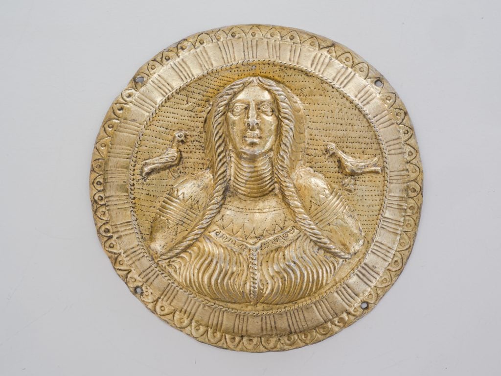 An circular silver bust of a goddess depicted with long plaits and two birds perched above her shoulders