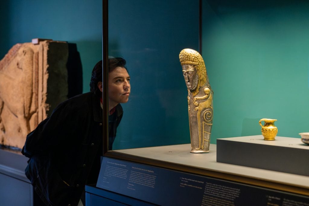 A man looking at an ancient gold sculpture that is displayed in a glass case in a gallery