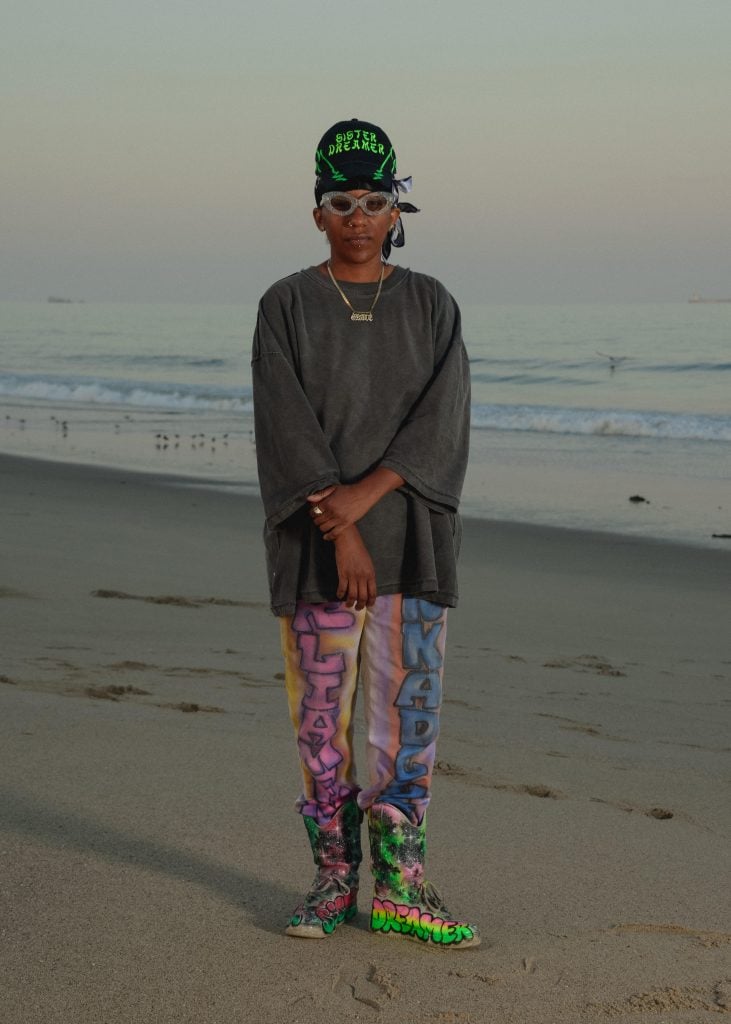 a woman in colorful pants, sunglasses and an oversized sweatshirt and baseball cap is on the beach last dusk