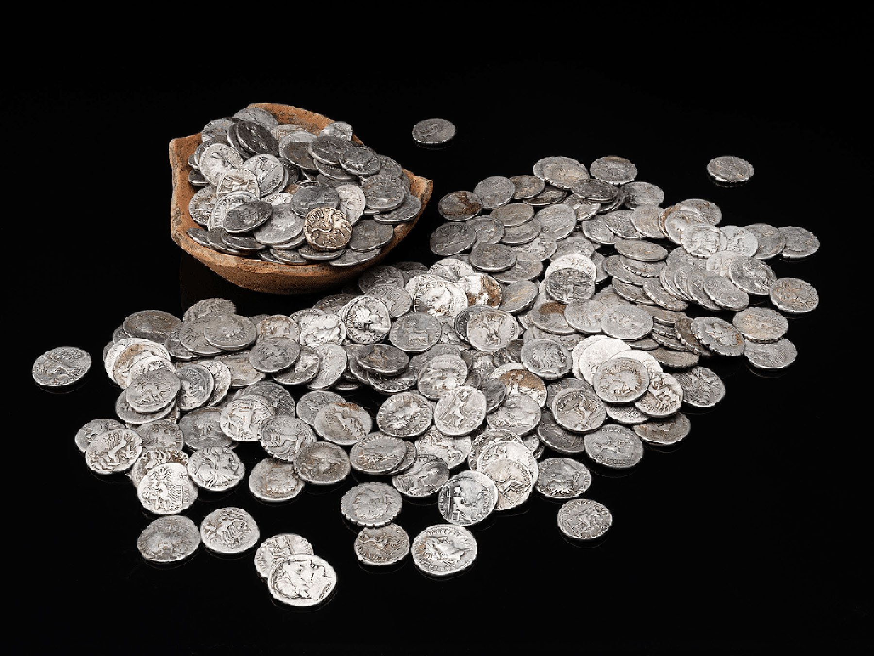 A trove of ancient Roman coins spilling off a bowl onto a black surface