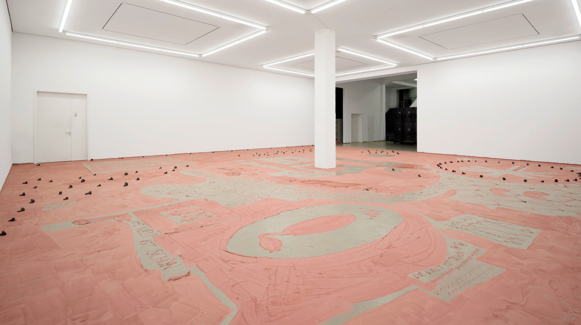 a large white walled room in a museum / gallery setting but the floor is covered in a layer of pink clay with markings, drawings, and text inscribed onto its surface.