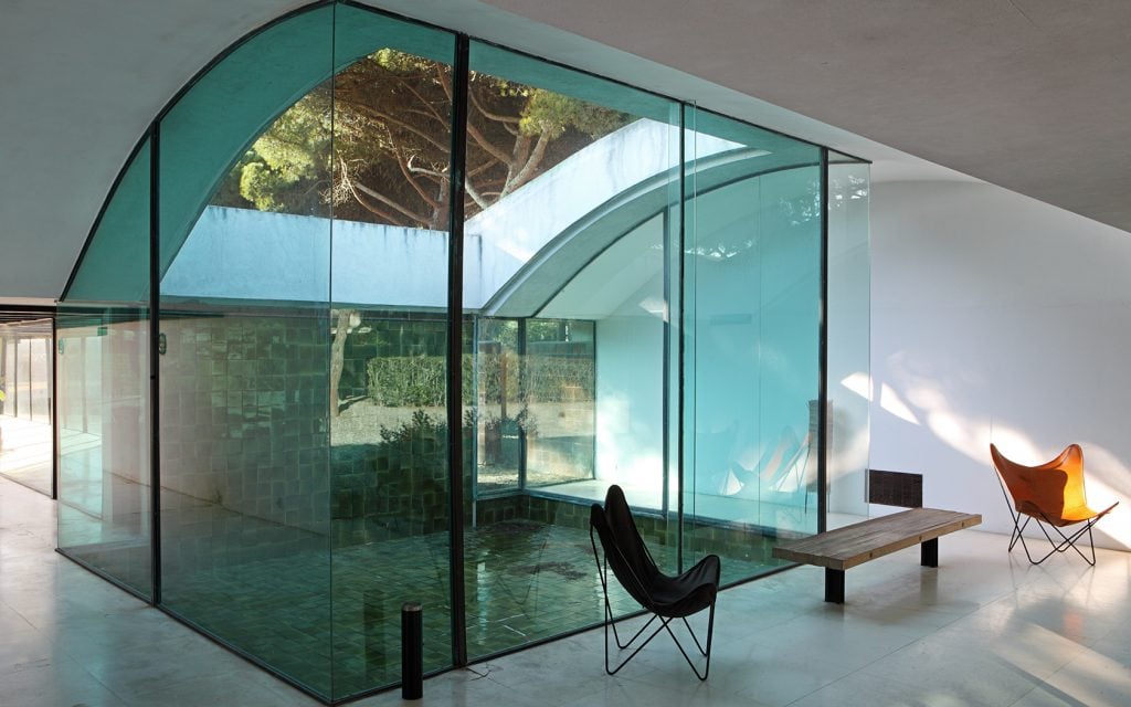 Sunlit interior of Casa Gomis featuring curved glass walls, green-tiled reflections, and modern butterfly chairs.